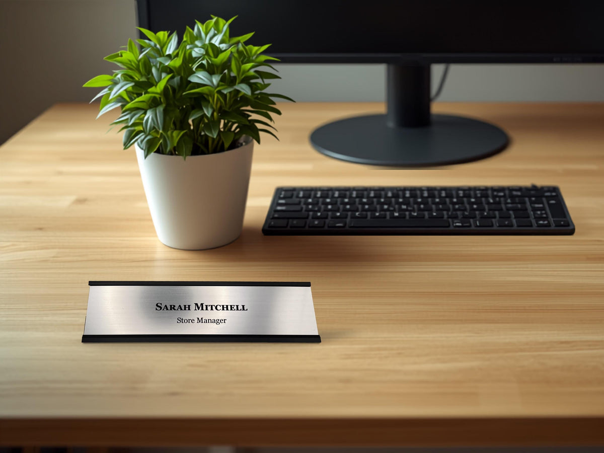A rectangular desk nameplate with a brushed silver metal-look face and black trim. The text "Sarah Mitchell" is engraved in a black serif font on the top line, with "Store Manager" in a smaller black font on the line below.