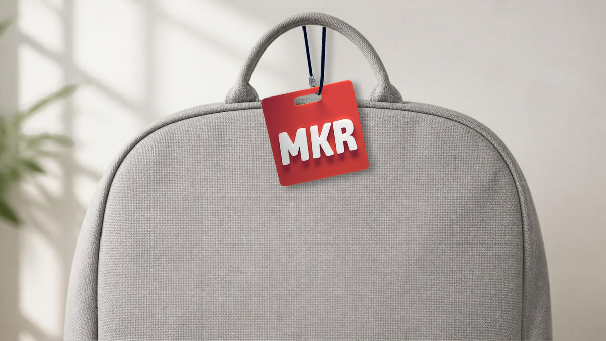 A coral bag tag with the initials MKR in raised white letters, hanging from its wire loop on the handle of a gray backpack.