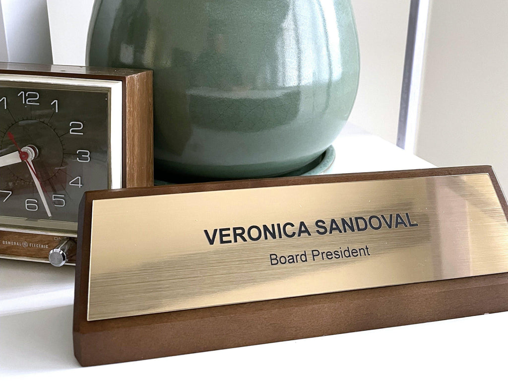 A desk nameplate with a brushed gold metal-look plate set into a dark walnut base. The text reads VERONICA SANDOVAL and Board President in black engraved lettering. A square analog clock and a green ceramic pot are visible in the background.