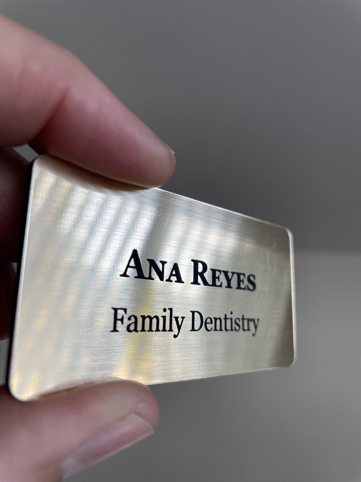 A rectangular name badge with a brushed gold metal-look finish. The text "ANA REYES" is engraved in a bold, black serif font on the top line, with "Family Dentistry" in a smaller black serif font on the line below.