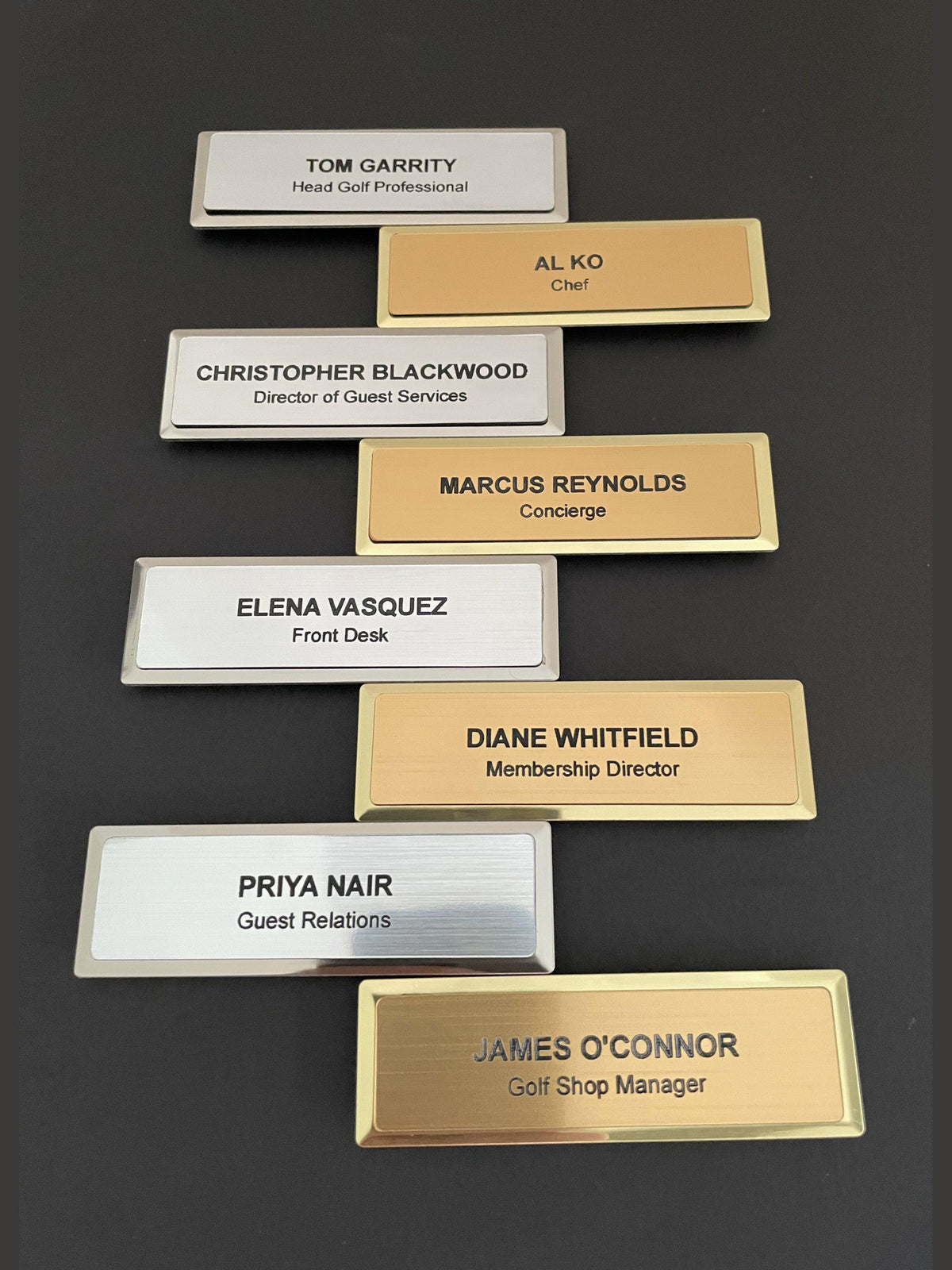 A collection of eight rectangular name badges arranged in a staggered pattern. Four badges feature a silver-toned finish with black text, and four feature a gold-toned finish with black text, each displaying a name and job title.