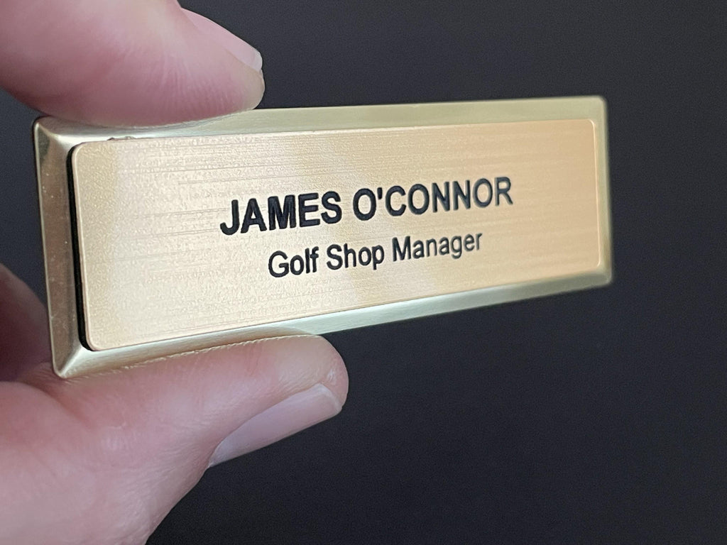 A name badge on a gold metal bar with a brushed metal-look face. The black engraved text reads JAMES O'CONNOR on the top line and Golf Shop Manager on the bottom line in a clean, sans-serif font.