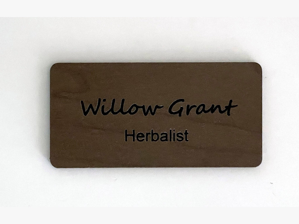 A rectangular brown wooden name tag with rounded corners. The text Willow Grant is engraved in a script font on the top line, and the word Herbalist is engraved in a simple sans-serif font on the bottom line of the wooden surface.