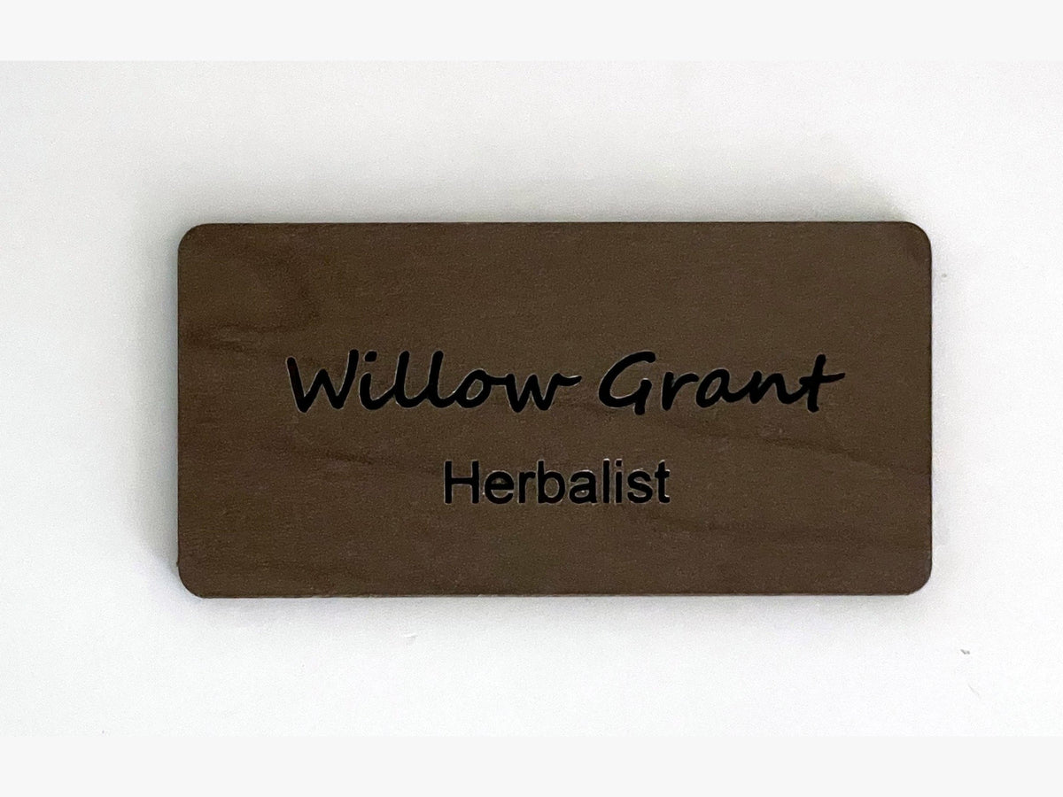 A rectangular brown wooden name tag with rounded corners. The text Willow Grant is engraved in a script font on the top line, and the word Herbalist is engraved in a simple sans-serif font on the bottom line of the wooden surface.