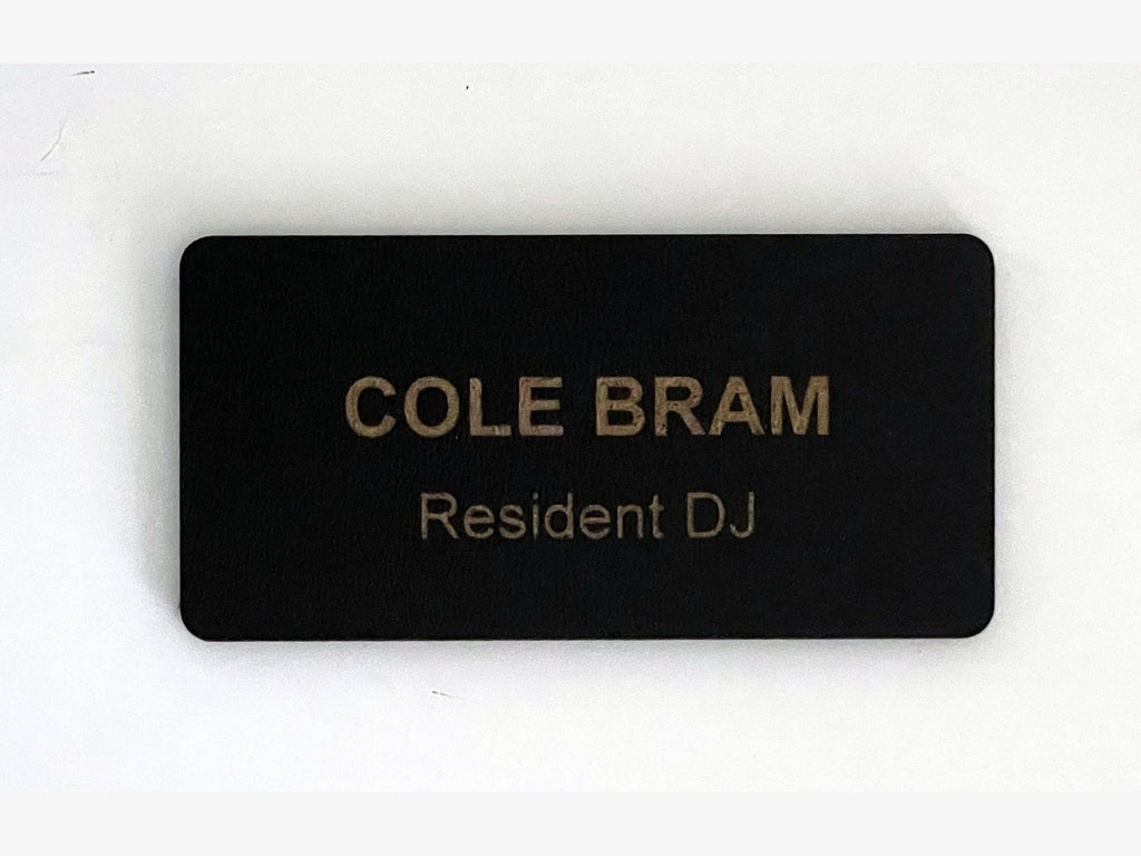 A rectangular black name badge with rounded corners featuring the engraved text COLE BRAM in large capital letters and the text Resident DJ in smaller letters below it. The surface has a matte finish and a consistent dark color.