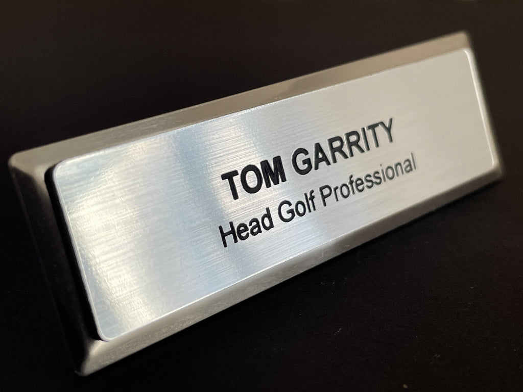 A name badge on a silver metal bar with a brushed metal-look face. The badge features the black engraved text TOM GARRITY on the top line and Head Golf Professional on the second line.