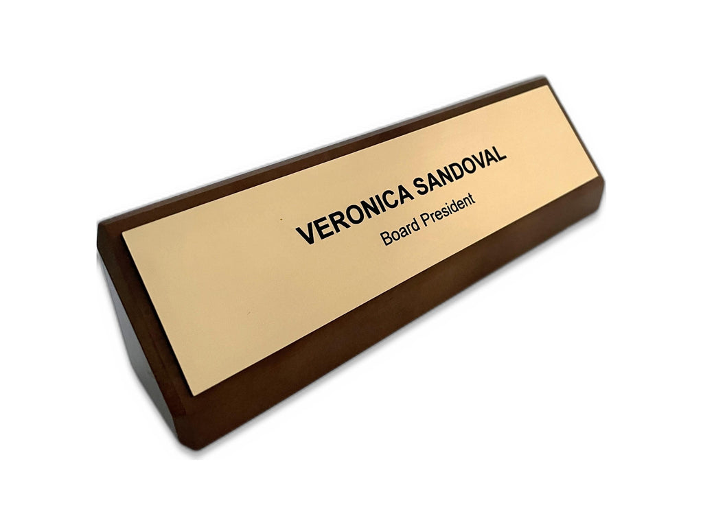 A triangular desk nameplate featuring a dark walnut base and a gold-toned, metal-look faceplate. The black engraved text reads VERONICA SANDOVAL followed by the title Board President on the line below in a sans-serif font.