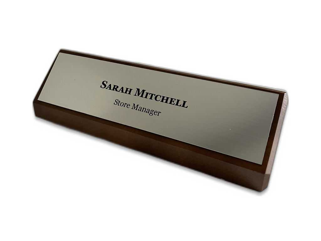 A rectangular desk nameplate featuring a dark walnut base and a silver metal-look faceplate. The text Sarah Mitchell is engraved in black, followed by the title Store Manager in a smaller black font centered on the plate.