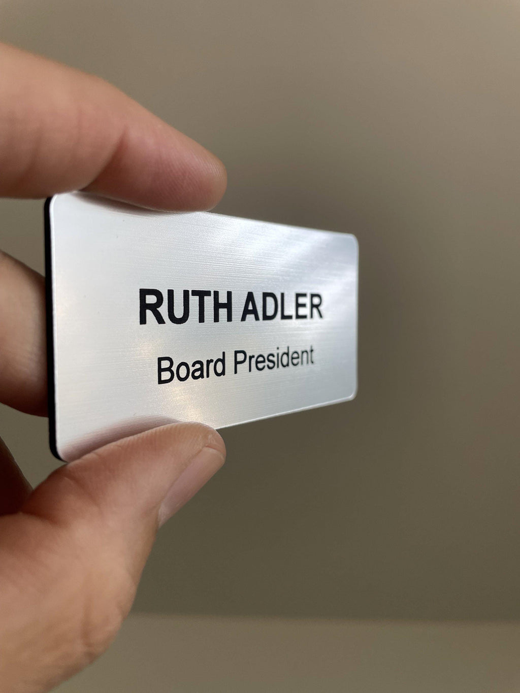 A rectangular silver-toned name badge with a brushed metal-look finish and black engraved text. The badge displays the name RUTH ADLER in large capital letters, with the title Board President in smaller black text directly below it.