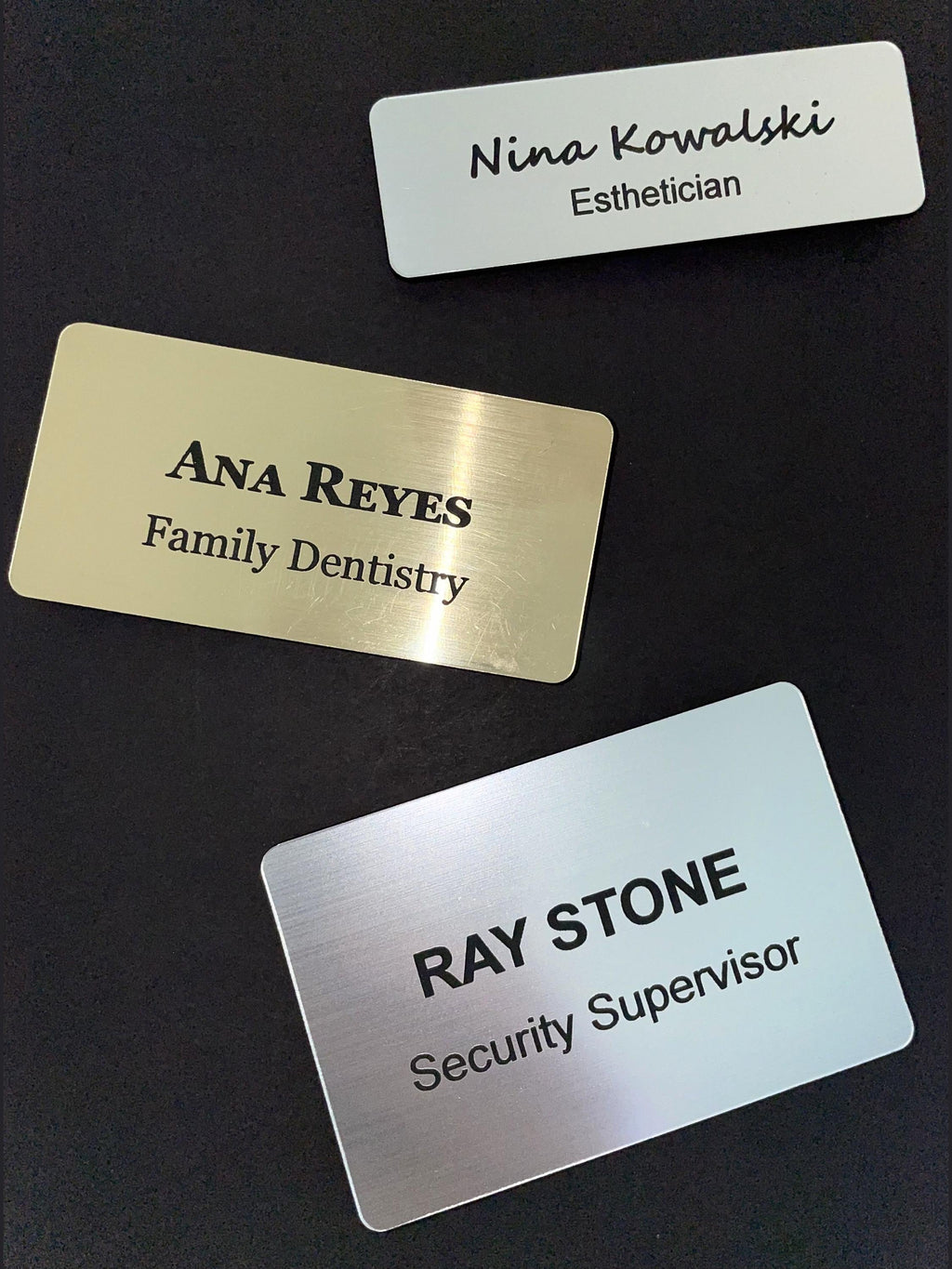 Three rectangular name badges with rounded corners. One silver badge reads Nina Kowalski Esthetician, one gold badge reads ANA REYES Family Dentistry, and one silver badge reads RAY STONE Security Supervisor in black text.