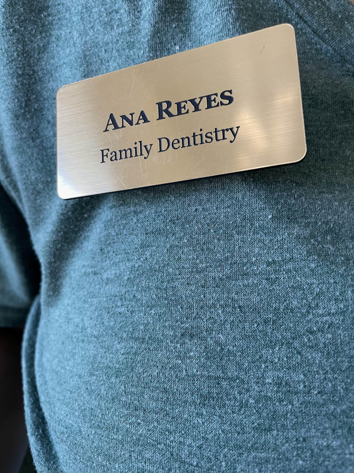A rectangular brushed silver-tone name badge with a metal-look finish and rounded corners. The badge features the engraved text "ANA REYES" in a bold serif font, followed by "Family Dentistry" in a smaller serif font.