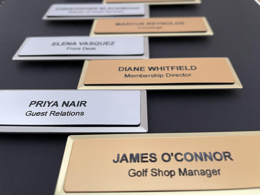 A collection of name badges on silver and gold metal bars with black engraved text. The badges feature names and job titles including James O'Connor, Priya Nair, Diane Whitfield, Elena Vasquez, and Marcus Reynolds.