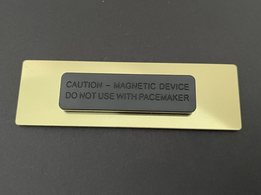 A rectangular gold-toned metal name badge backing featuring a centered black magnetic attachment. The black magnetic piece is embossed with the text: CAUTION - MAGNETIC DEVICE DO NOT USE WITH PACEMAKER.