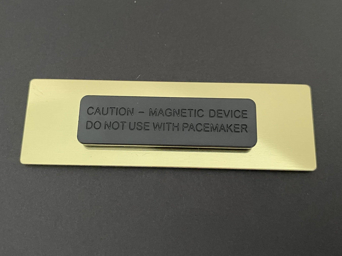 A rectangular gold-toned metal name badge backing featuring a centered black magnetic attachment. The black magnetic piece is embossed with the text: CAUTION - MAGNETIC DEVICE DO NOT USE WITH PACEMAKER.