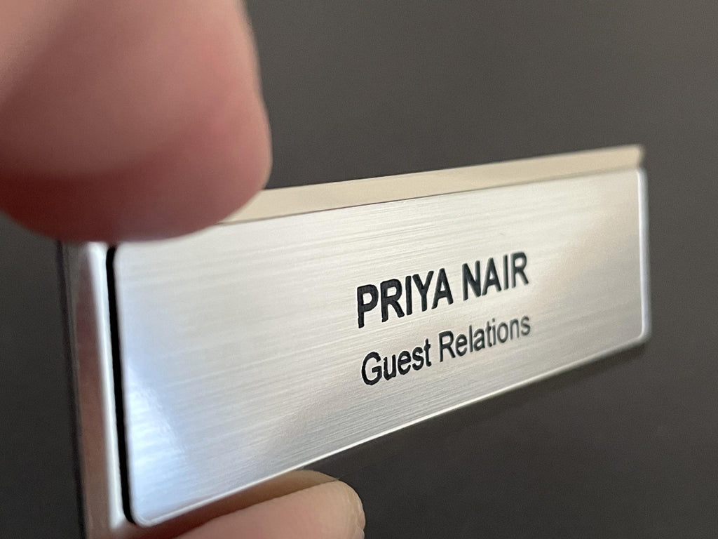 A name badge on a silver metal bar with a brushed metal-look face. The badge features the engraved text PRIYA NAIR in a bold, uppercase sans-serif font, followed by the text Guest Relations in a smaller, standard sans-serif font.