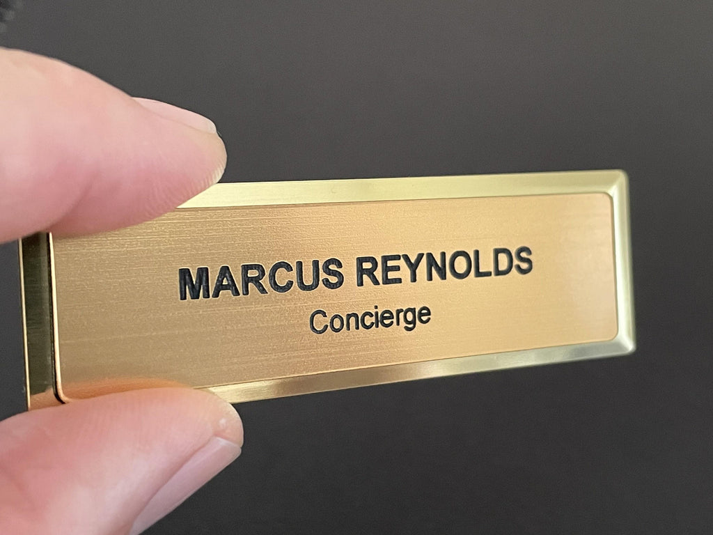 A name badge on a gold metal bar with a brushed metal-look face. The badge features the black engraved text MARCUS REYNOLDS on the top line and the word Concierge on the line below in a smaller font size.