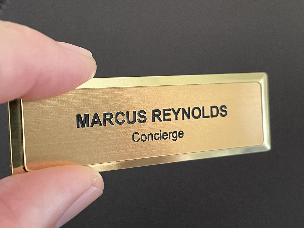 A name badge on a gold metal bar with a brushed metal-look face. The badge features the black engraved text MARCUS REYNOLDS on the top line and the word Concierge on the line below in a smaller font size.