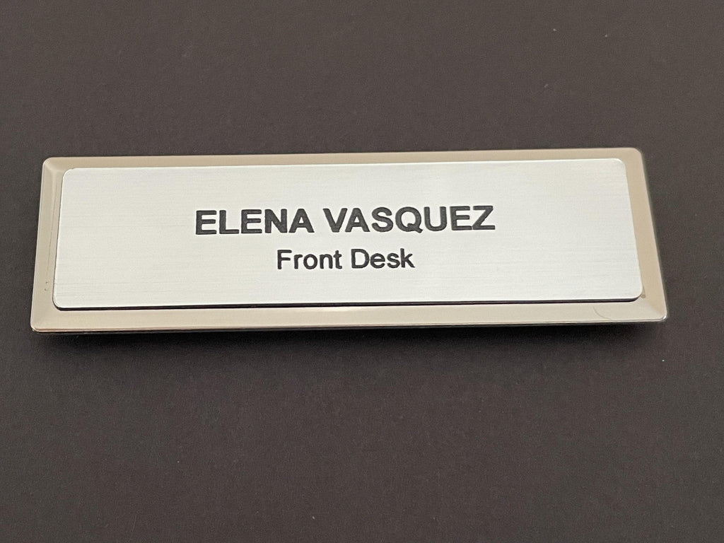 A name badge on a silver metal bar with a brushed metal-look face. The text ELENA VASQUEZ is engraved in bold black capital letters, with the words Front Desk in smaller black letters underneath.