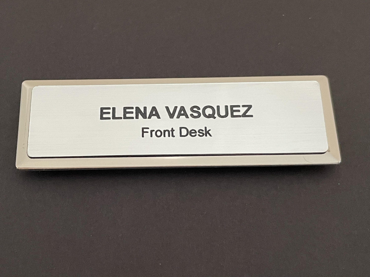 A name badge on a silver metal bar with a brushed metal-look face. The text ELENA VASQUEZ is engraved in bold black capital letters, with the words Front Desk in smaller black letters underneath.