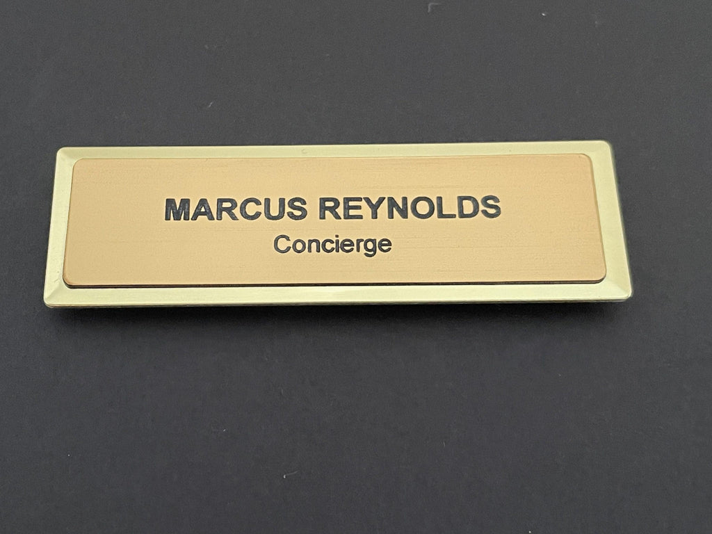 A name badge on a gold-colored metal bar with a brushed gold-tone, metal-look face. The text MARCUS REYNOLDS is engraved in black capital letters on the top line, with the word Concierge below in black.