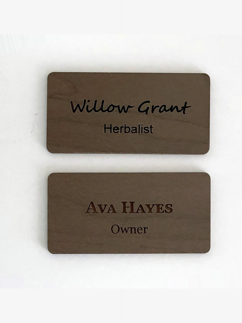 Two rectangular brown wooden name badges with rounded corners. The top badge is engraved with the text Willow Grant and Herbalist. The bottom badge is engraved with the text AVA HAYES and Owner in a different font style.