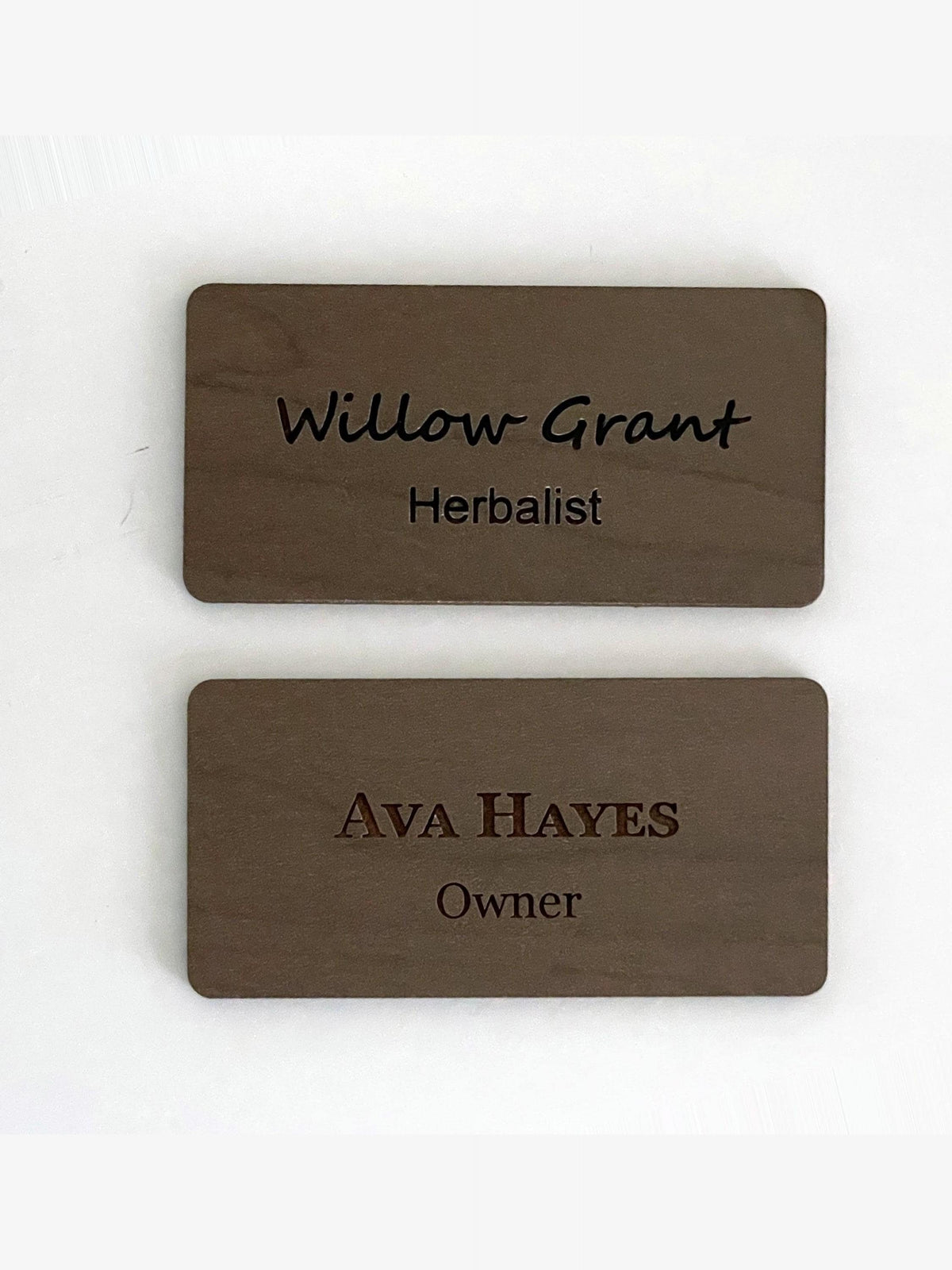 Two rectangular brown wooden name badges with rounded corners. The top badge is engraved with the text Willow Grant and Herbalist. The bottom badge is engraved with the text AVA HAYES and Owner in a different font style.