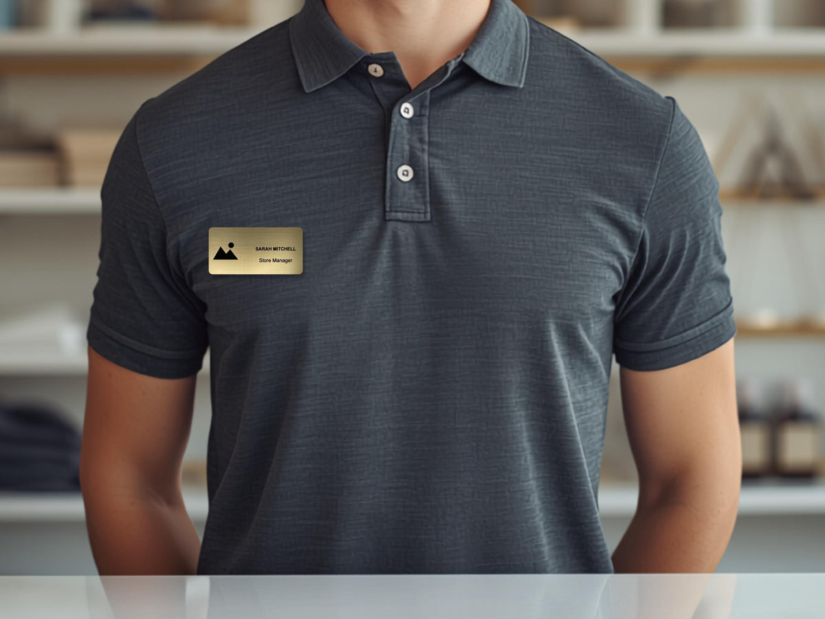 A dark gray short-sleeved polo shirt with a three-button placket. Attached to the chest is a rectangular gold-toned name badge featuring a small black mountain icon, the name Sarah Mitchell, and the job title Store Manager.