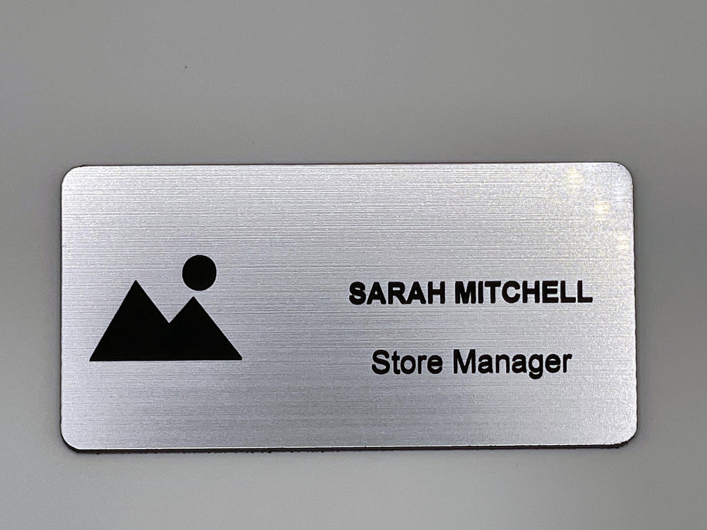 A rectangular silver-toned name badge with a brushed metal-look finish. The left side features a black graphic of a mountain and a sun. The right side displays the text SARAH MITCHELL in uppercase letters, followed by the text Store Manager.