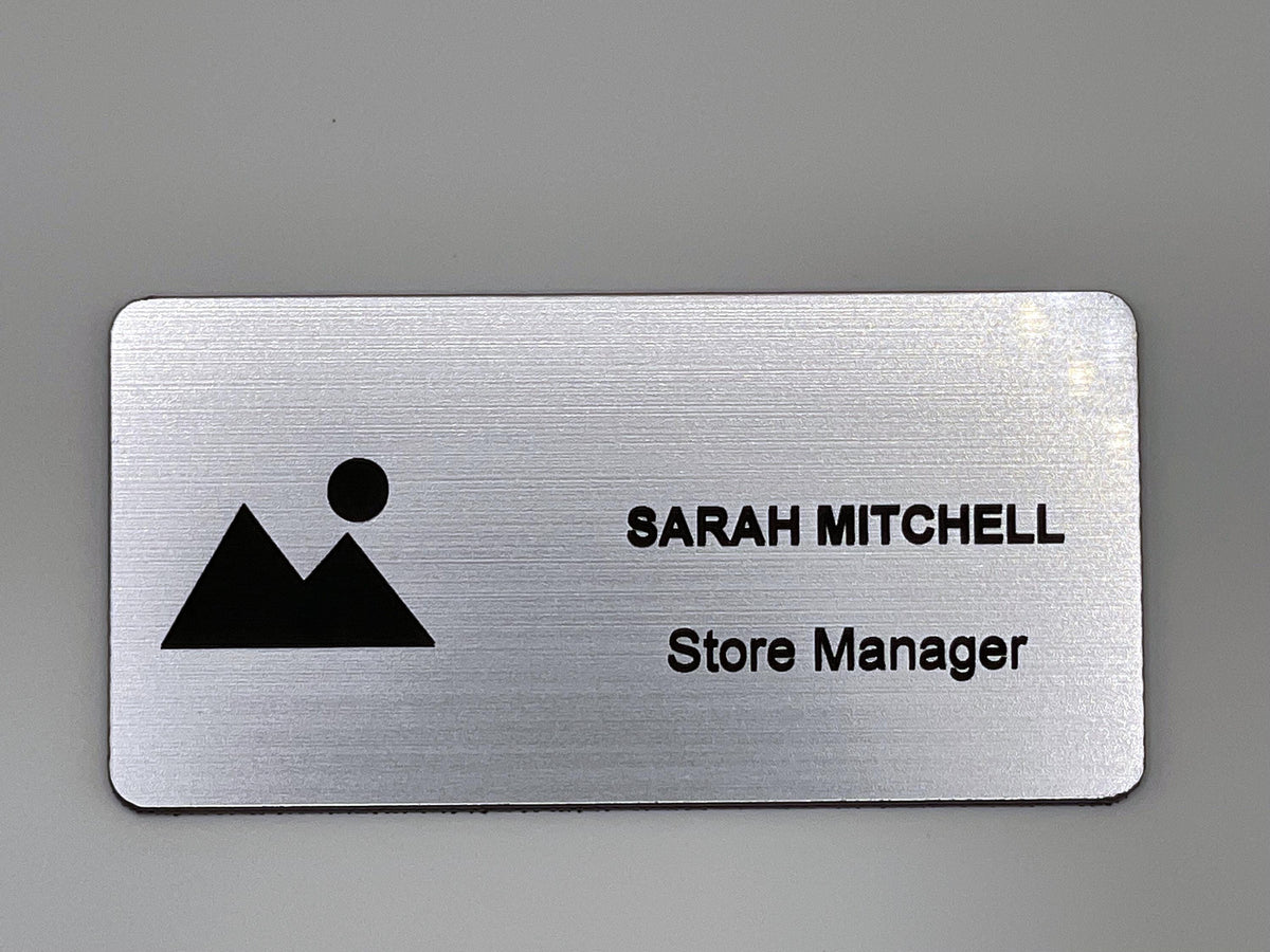 A rectangular silver-toned name badge with a brushed metal-look finish. The left side features a black graphic of a mountain and a sun. The right side displays the text SARAH MITCHELL in uppercase letters, followed by the text Store Manager.