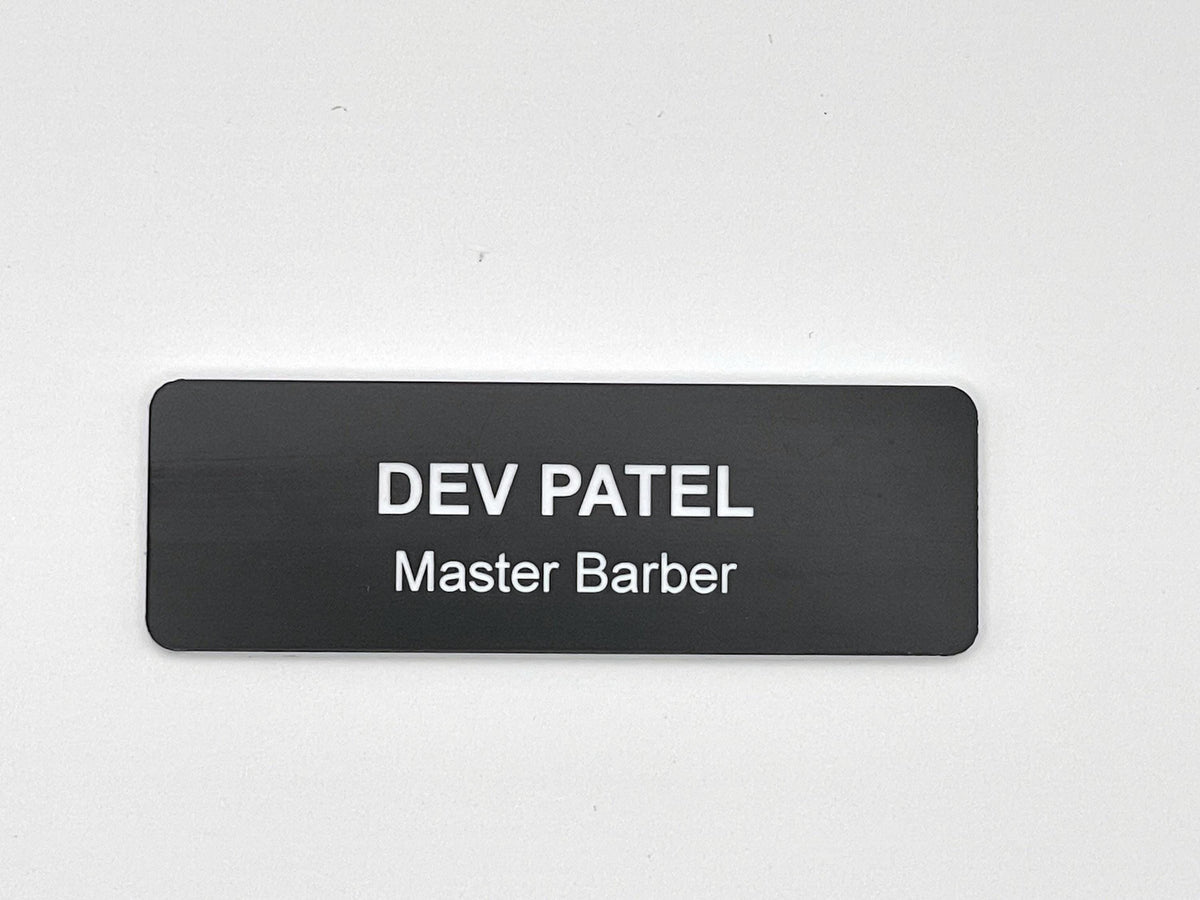 A rectangular black name badge with rounded corners featuring the white text DEV PATEL on the top line and Master Barber on the bottom line in a clean sans-serif font, centered against the dark background.
