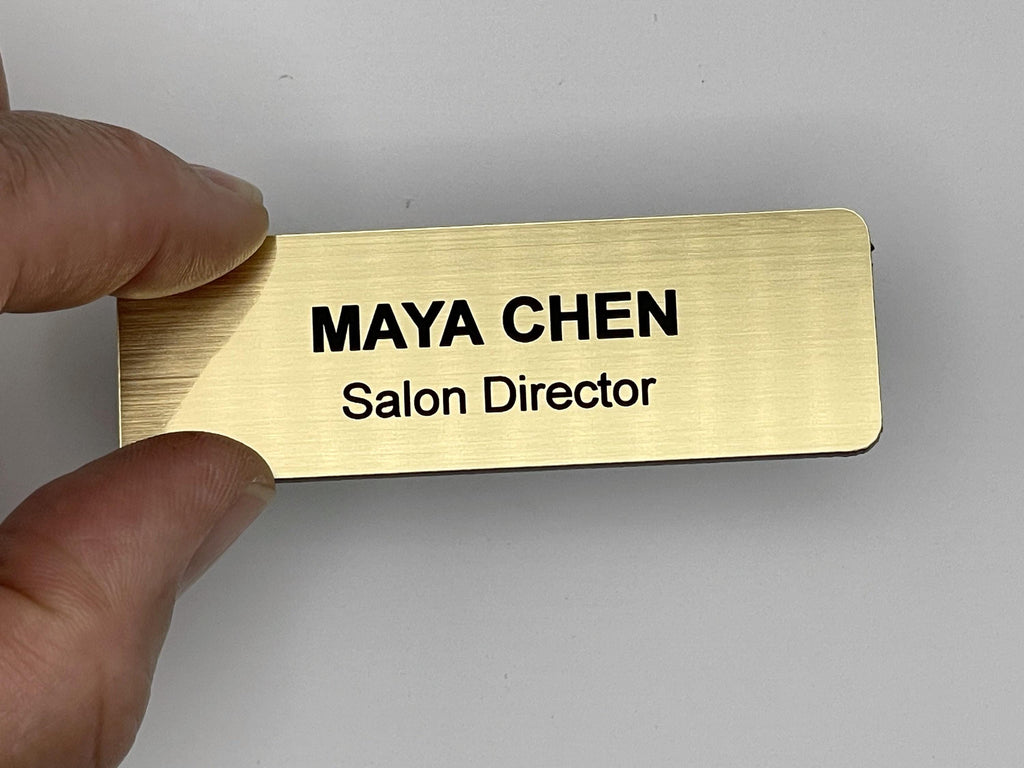 A rectangular gold-toned name badge with a brushed metal-look finish. The text "MAYA CHEN" is engraved in bold black capital letters on the top line, with "Salon Director" in smaller black letters on the line below.