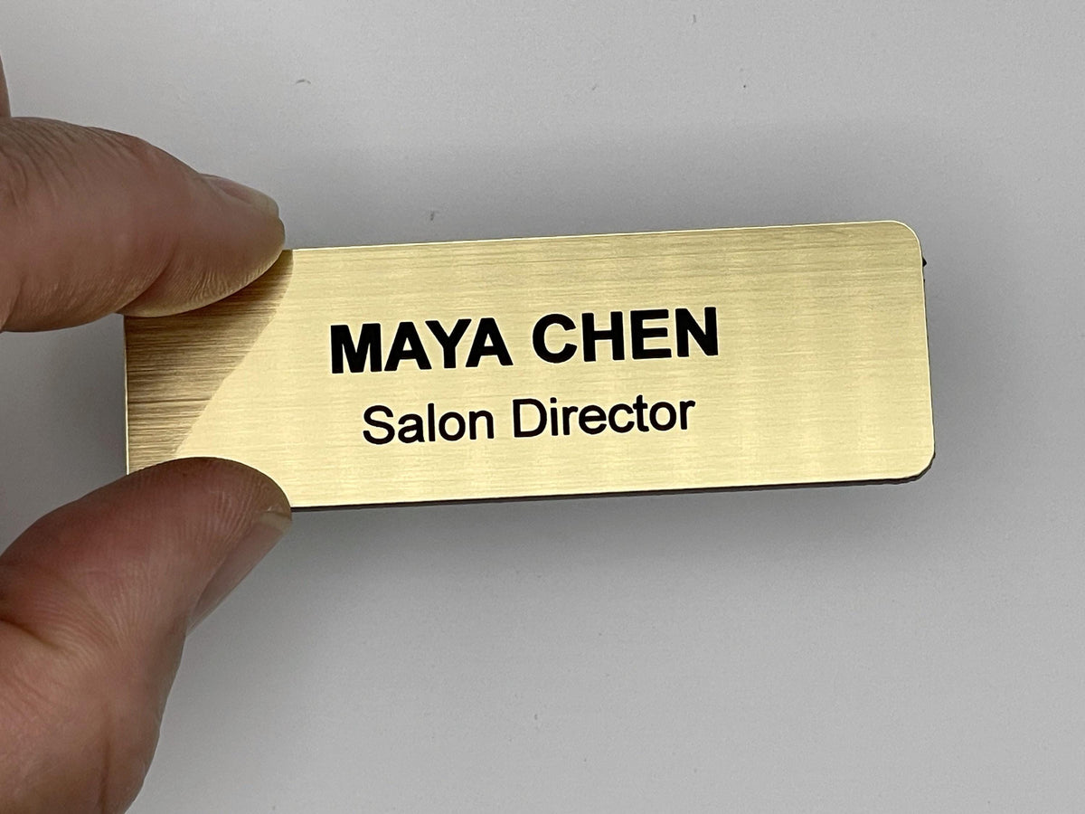 A rectangular gold-toned name badge with a brushed metal-look finish. The text "MAYA CHEN" is engraved in bold black capital letters on the top line, with "Salon Director" in smaller black letters on the line below.