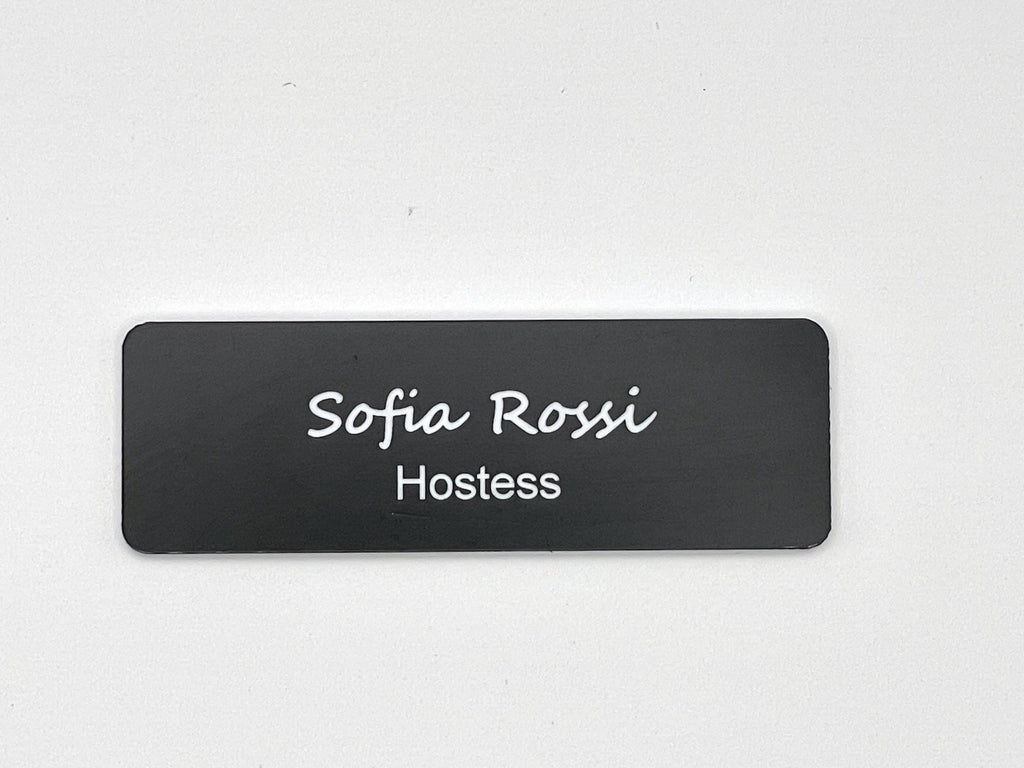A rectangular black name badge with rounded corners featuring the white text Sofia Rossi in a script font on the top line and the word Hostess in a clean sans-serif font on the bottom line, centered on the badge surface.