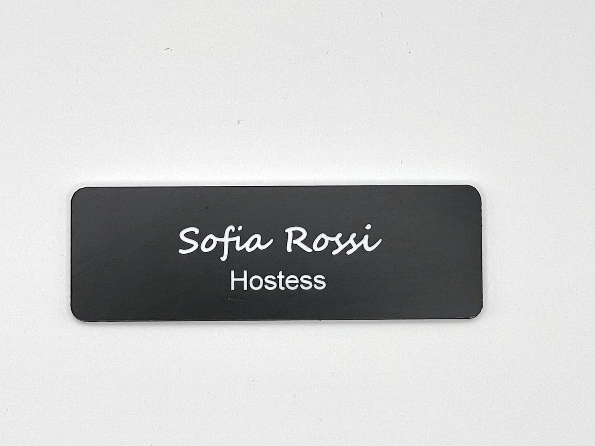 A rectangular black name badge with rounded corners featuring the white text Sofia Rossi in a script font on the top line and the word Hostess in a clean sans-serif font on the bottom line, centered on the badge surface.