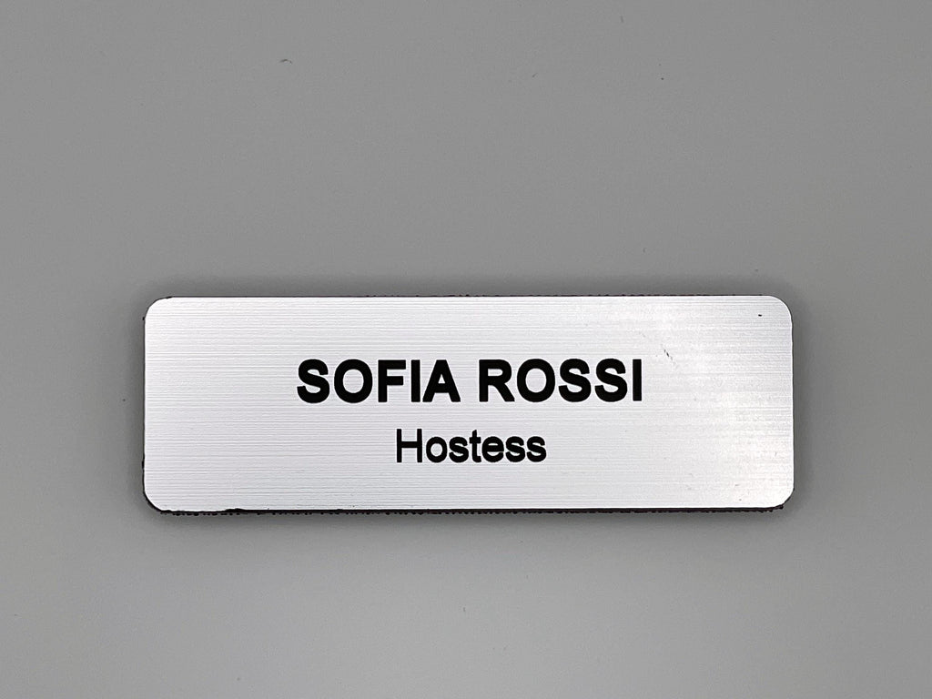 A rectangular silver-toned name badge with a brushed metal-look finish. The text SOFIA ROSSI is engraved in bold black capital letters on the top line, with the word Hostess in smaller black letters directly underneath it.