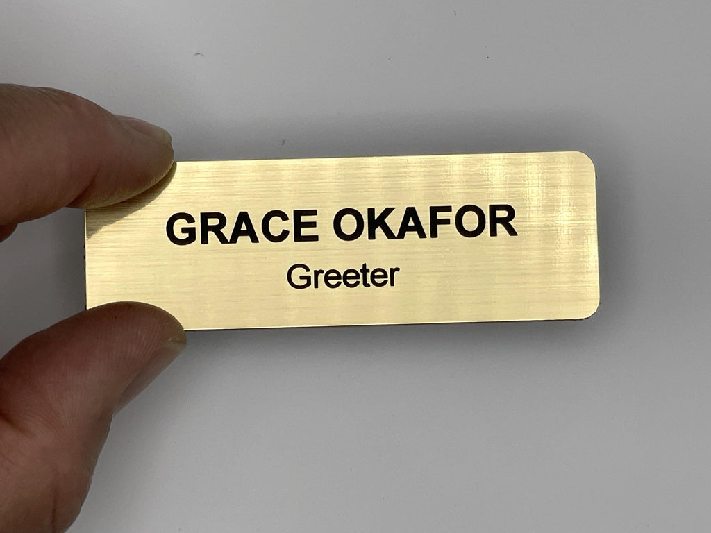 A rectangular gold-toned name badge with a brushed metal-look finish. The badge features the name GRACE OKAFOR engraved in bold black capital letters, with the job title Greeter in smaller black letters directly underneath the name.