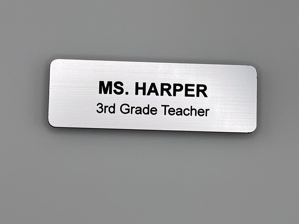 A rectangular silver-toned name badge with a brushed metal-look finish. The text is engraved in black, featuring the name MS. HARPER on the top line and the job title 3rd Grade Teacher on the bottom line. The badge has rounded corners.