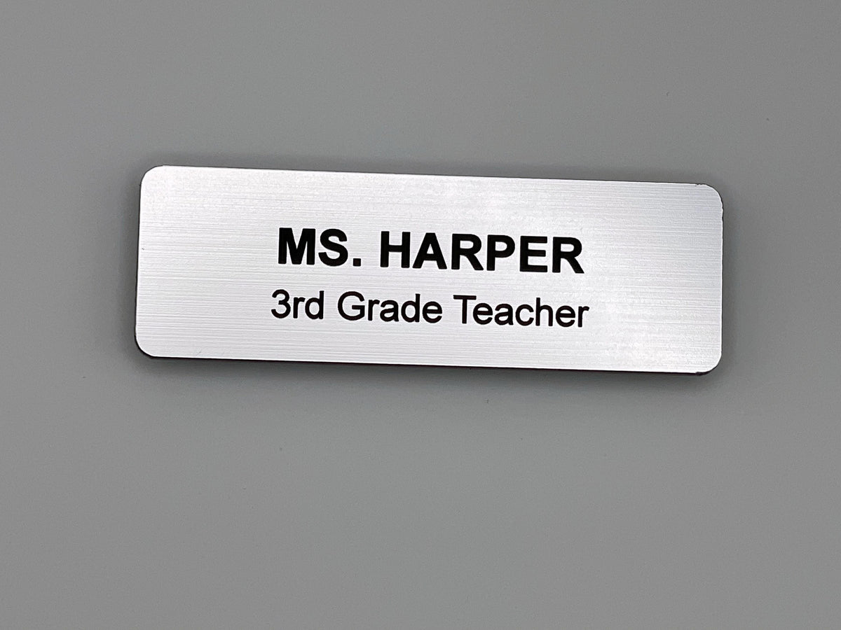 A rectangular silver-toned name badge with a brushed metal-look finish. The text is engraved in black, featuring the name MS. HARPER on the top line and the job title 3rd Grade Teacher on the bottom line. The badge has rounded corners.