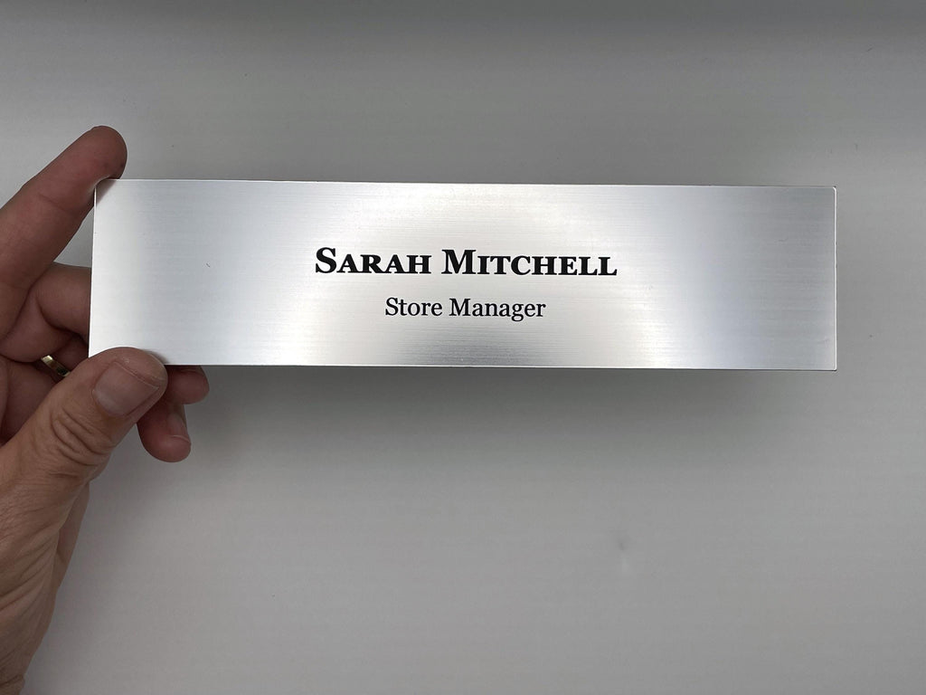 A rectangular silver-toned nameplate with a brushed metal-look finish. The text "SARAH MITCHELL" is engraved in a bold, black serif font on the top line, with "Store Manager" in a smaller, black sans-serif font below it.