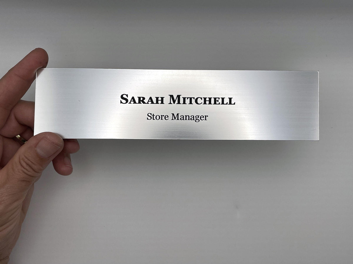 A rectangular silver-toned nameplate with a brushed metal-look finish. The text "SARAH MITCHELL" is engraved in a bold, black serif font on the top line, with "Store Manager" in a smaller, black sans-serif font below it.