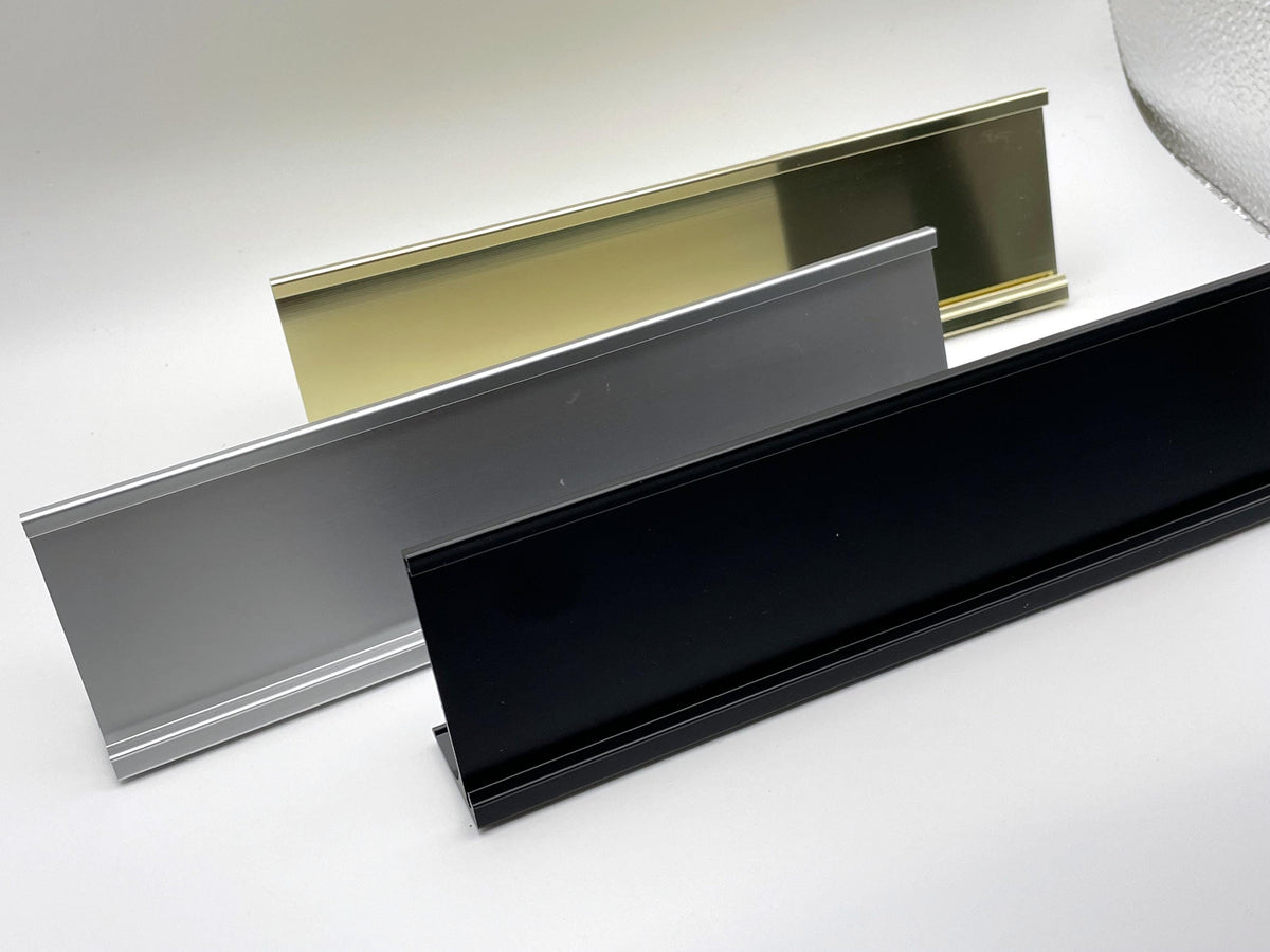 Three rectangular desk nameplate holders arranged in a staggered formation. The holders feature a smooth, angled front face with a top lip for inserting the nameplate. The set includes one gold-toned, one silver-toned, and one matte black finish.