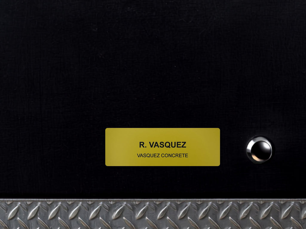 A black surface featuring a rectangular yellow nameplate with the text R. VASQUEZ and VASQUEZ CONCRETE engraved in black. To the right of the nameplate is a small, circular silver button. A silver diamond plate border runs along the bottom.