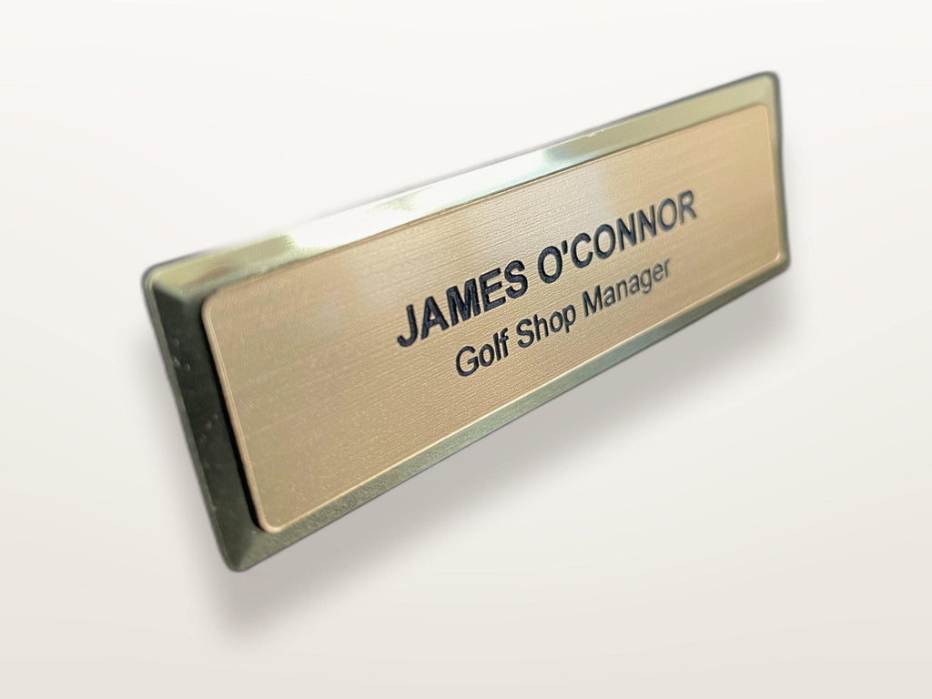 A name badge with a brushed gold-tone, metal-look face on a metal bar. The text JAMES O'CONNOR is engraved in black capital letters, followed by the title Golf Shop Manager in smaller black lettering.