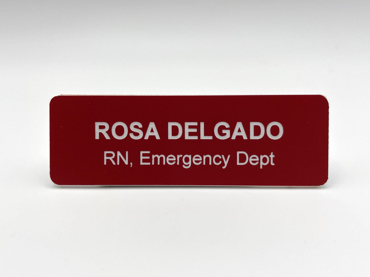 A rectangular name badge with a deep red background and white text. The name ROSA DELGADO is engraved in large capital letters on the top line, followed by the text RN, Emergency Dept on the second line in a smaller font size.