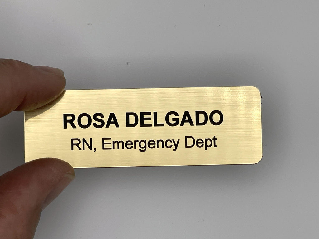 A rectangular gold-toned name badge with a brushed metal-look finish. The engraved black text reads ROSA DELGADO on the first line and RN, Emergency Dept on the second line. The badge has rounded corners and a smooth, metal-look surface.