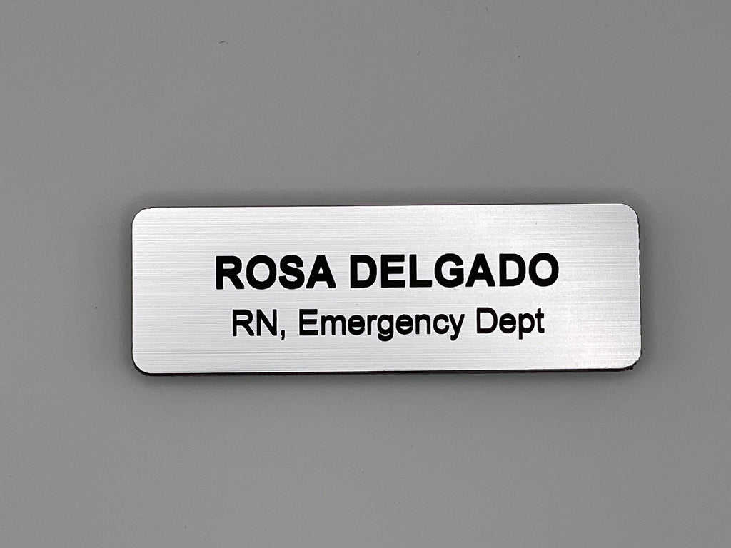 A rectangular silver-toned name badge with a brushed metal-look finish. The engraved black text reads ROSA DELGADO on the top line and RN, Emergency Dept on the bottom line. The badge features rounded corners and a clean design.