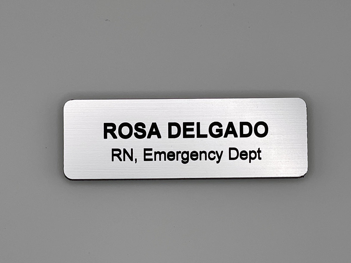 A rectangular silver-toned name badge with a brushed metal-look finish. The engraved black text reads ROSA DELGADO on the top line and RN, Emergency Dept on the bottom line. The badge features rounded corners and a clean design.