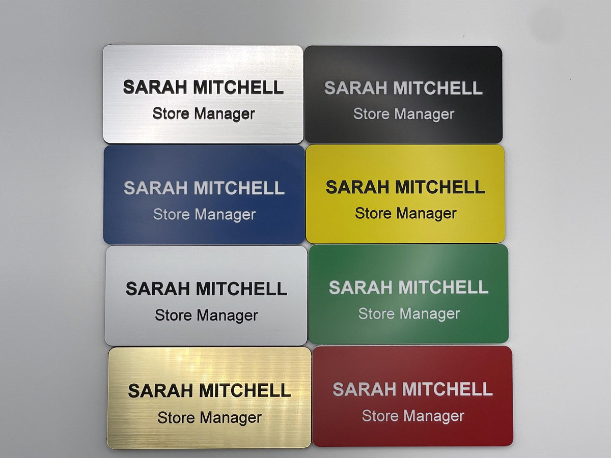 A collection of eight rectangular name badges in silver, black, blue, yellow, white, green, gold, and red. Each badge features the engraved text SARAH MITCHELL and Store Manager in a clean, sans-serif font with contrasting colors.