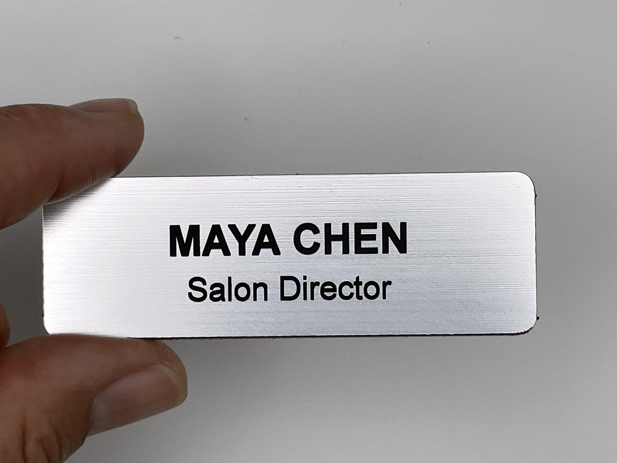 A rectangular silver-toned name badge with a brushed metal-look finish. The text is engraved in black, featuring the name MAYA CHEN in a bold, uppercase font on the top line and the title Salon Director on the bottom line.