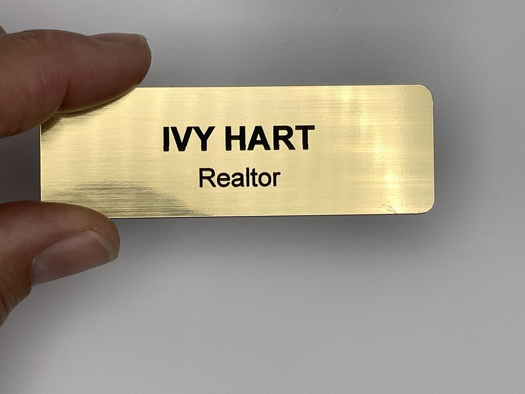 A rectangular gold-colored name badge with a brushed metal-look finish. The text IVY HART is engraved in bold black capital letters on the top line, with the word Realtor in smaller black letters directly underneath the name.